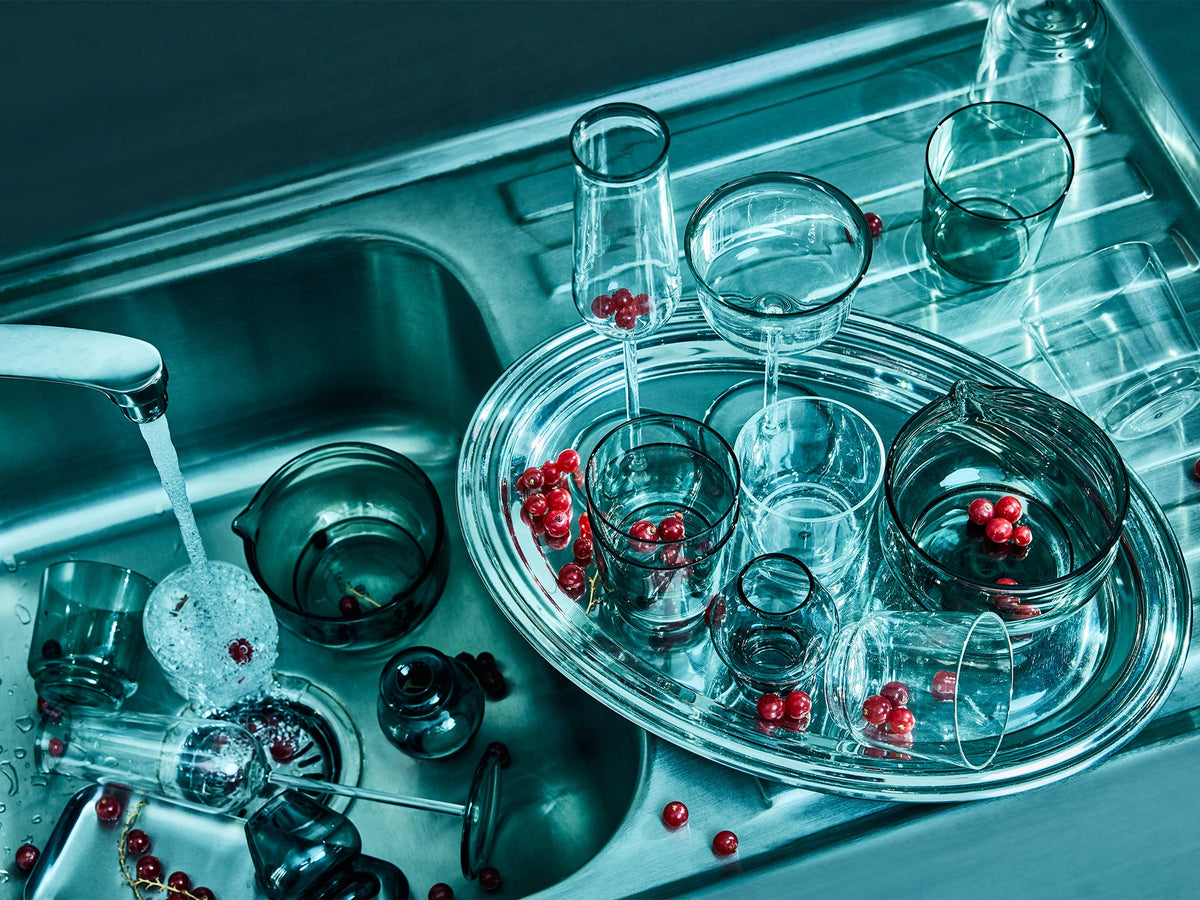 A collection of clear glass drinking vessels and a silver oval tray filled with small red berries, arranged in a stainless steel sink with running water.