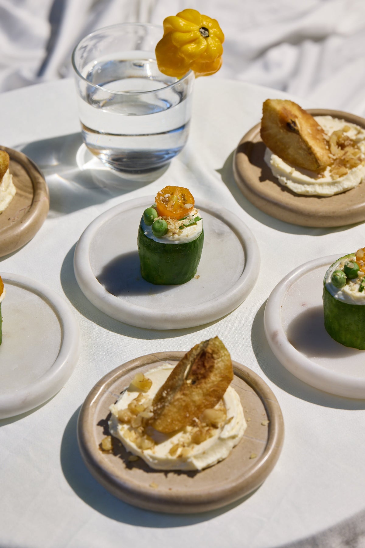 A styled food spread on a white tablecloth with marble coasters, featuring small appetizers including cucumber wraps with cream and orange topping, and topped with crispy fried strips, alongside a glass of water and a yellow chilli garnish.