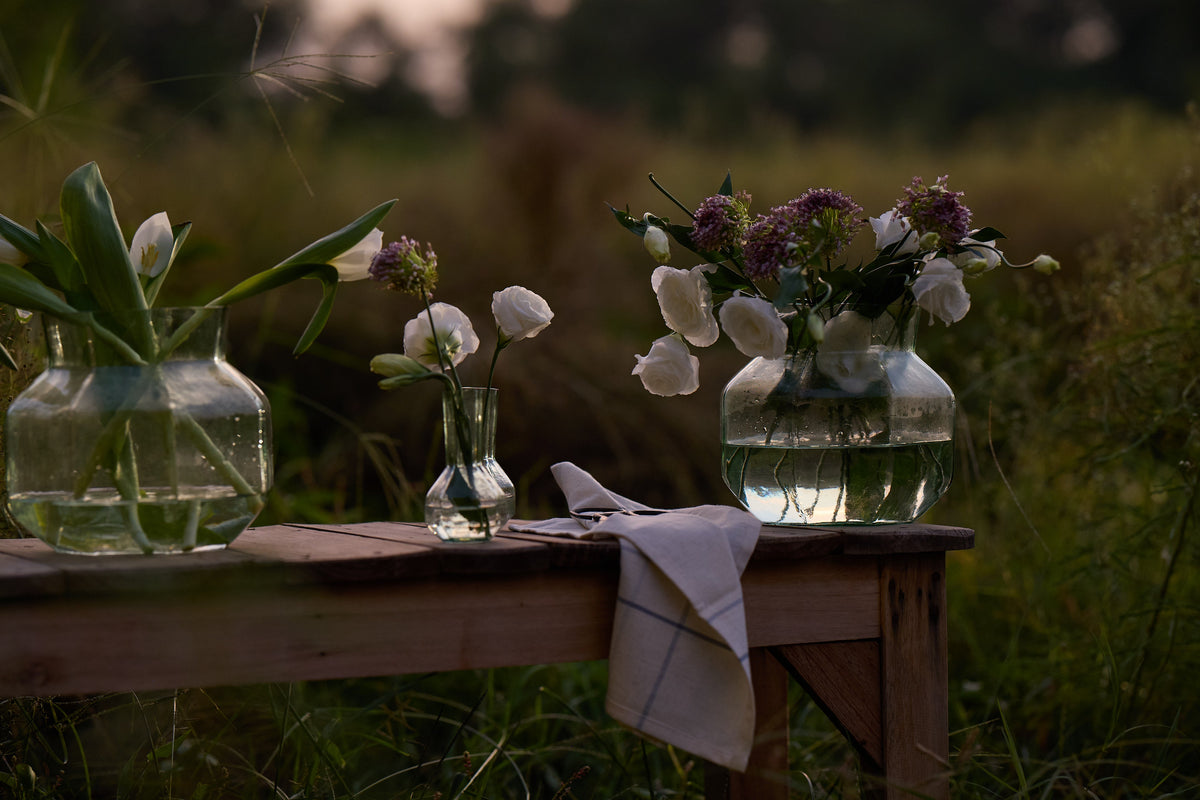 Three clear glass vases with water and flowers arranged on a wooden bench outdoors in a grassy field at dusk.