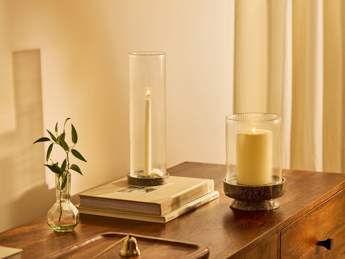 Two tall glass candle holders with white candles displayed on a wooden dresser alongside a small green plant in a glass vase, under warm ambient light.