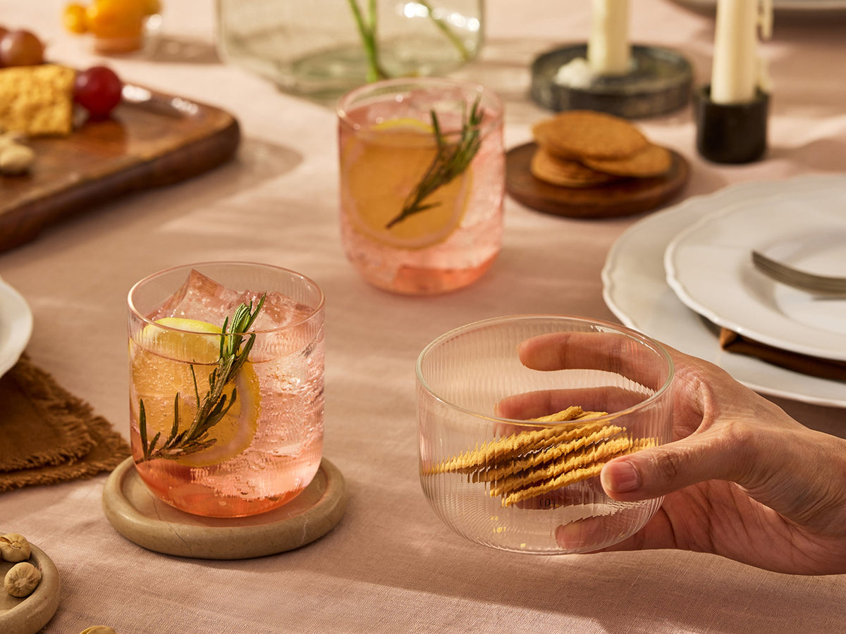 A hand holds a clear ribbed glass bowl containing crackers on a styled dining table. Two pink cocktails with lemon slices and rosemary garnish sit on wooden coasters in the background, with plates, candles, and food props around them.