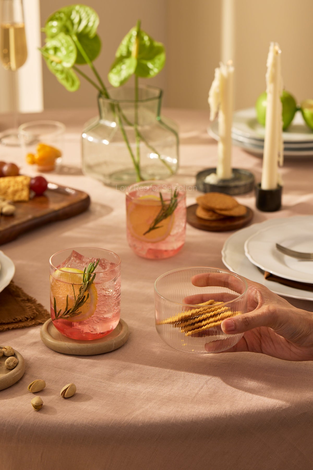 A hand holds a clear round ribbed glass tumbler with crackers inside, set on a pink tablecloth at a dinner table with cocktails, candles, plates, and green leaves in a vase.