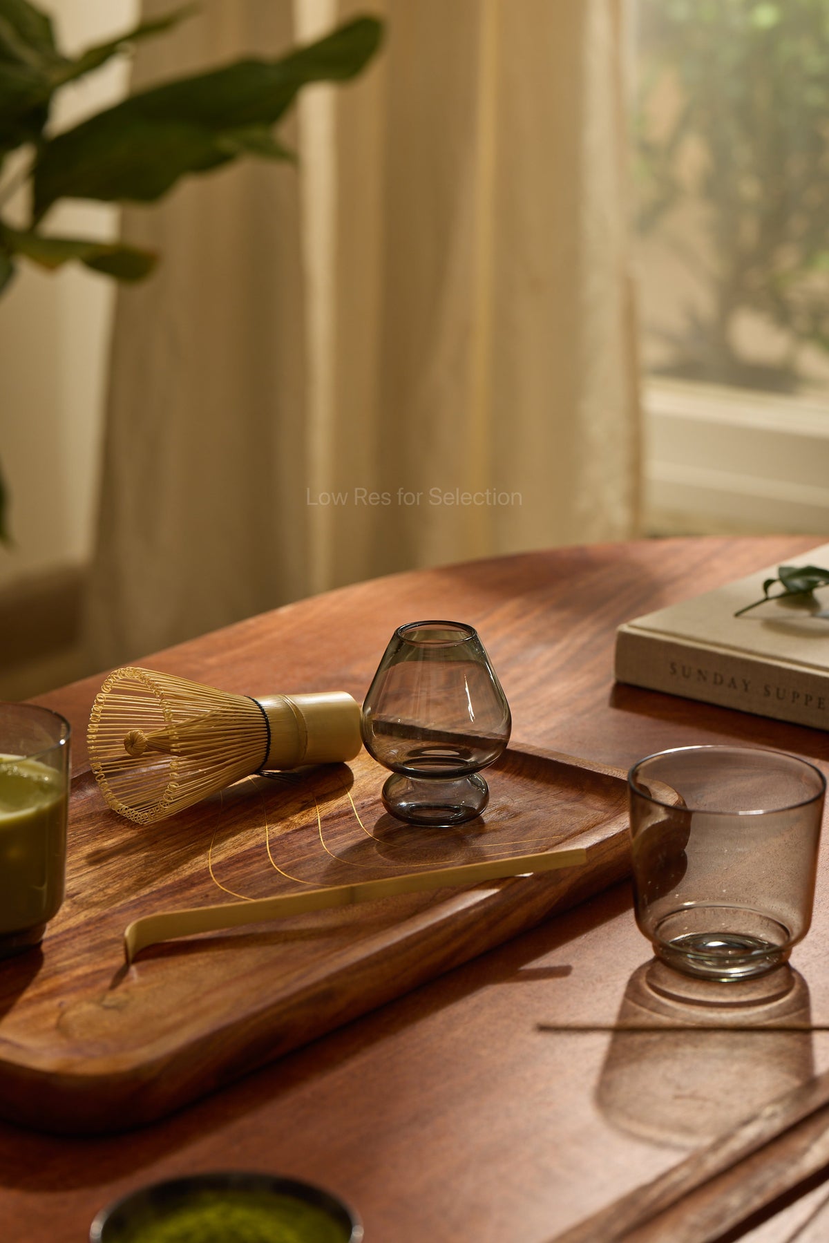 A styled tabletop scene with a wooden tray holding a bamboo whisk and grey stand, alongside two translucent drinking glasses on a warm wood table in a softly lit interior.