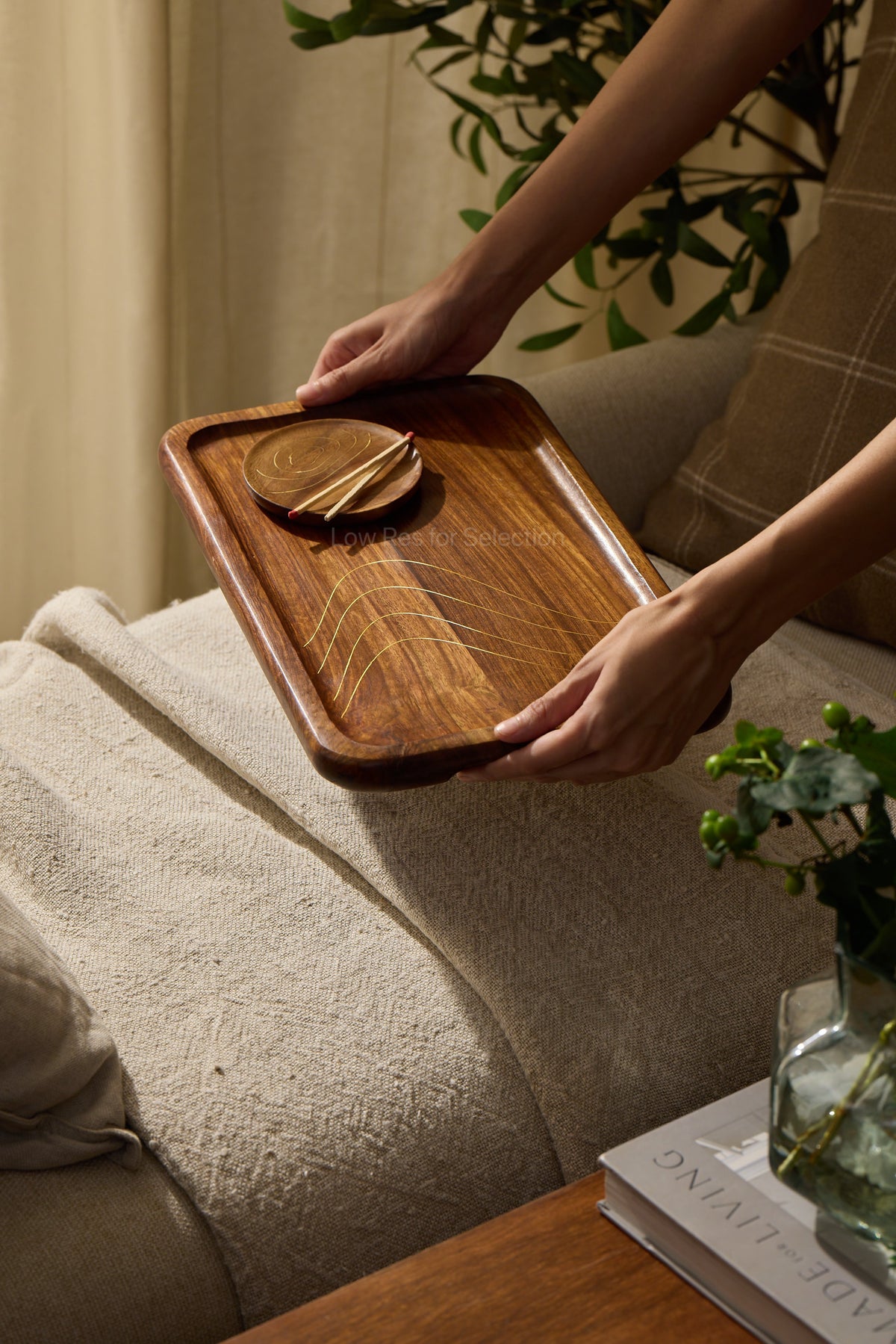 A person holds a rectangular wooden serving tray with rounded corners and brass inlay detail along with  a small matching wooden coaster on top, set in a cozy living room next to a sofa and a glass vase.