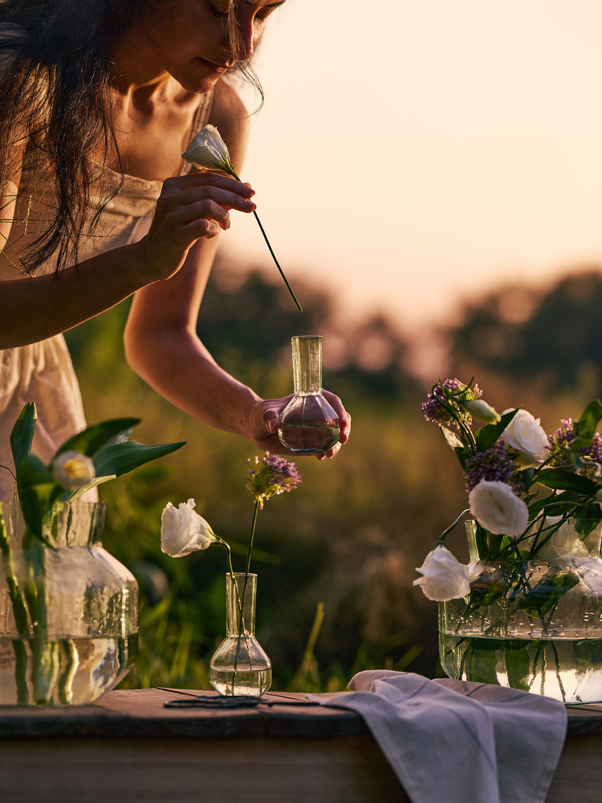 A woman in a light dress holds a small glass vase over a table set with flowers and glass vases at sunset. The scene is softly lit with warm golden light and a blurred outdoor background.