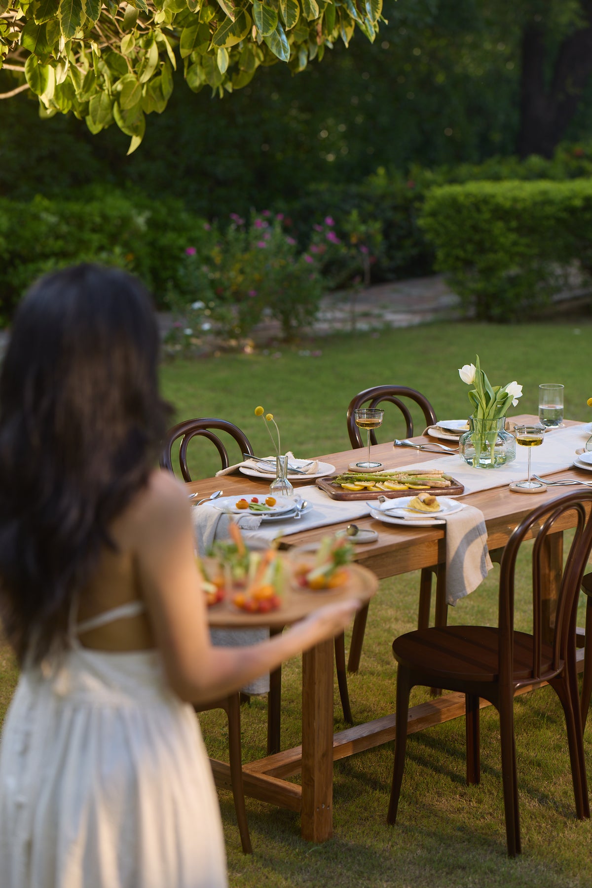 A woman in a white dress walks towards an outdoor dining table set with plates, glasses, and flowers in a lush garden setting.