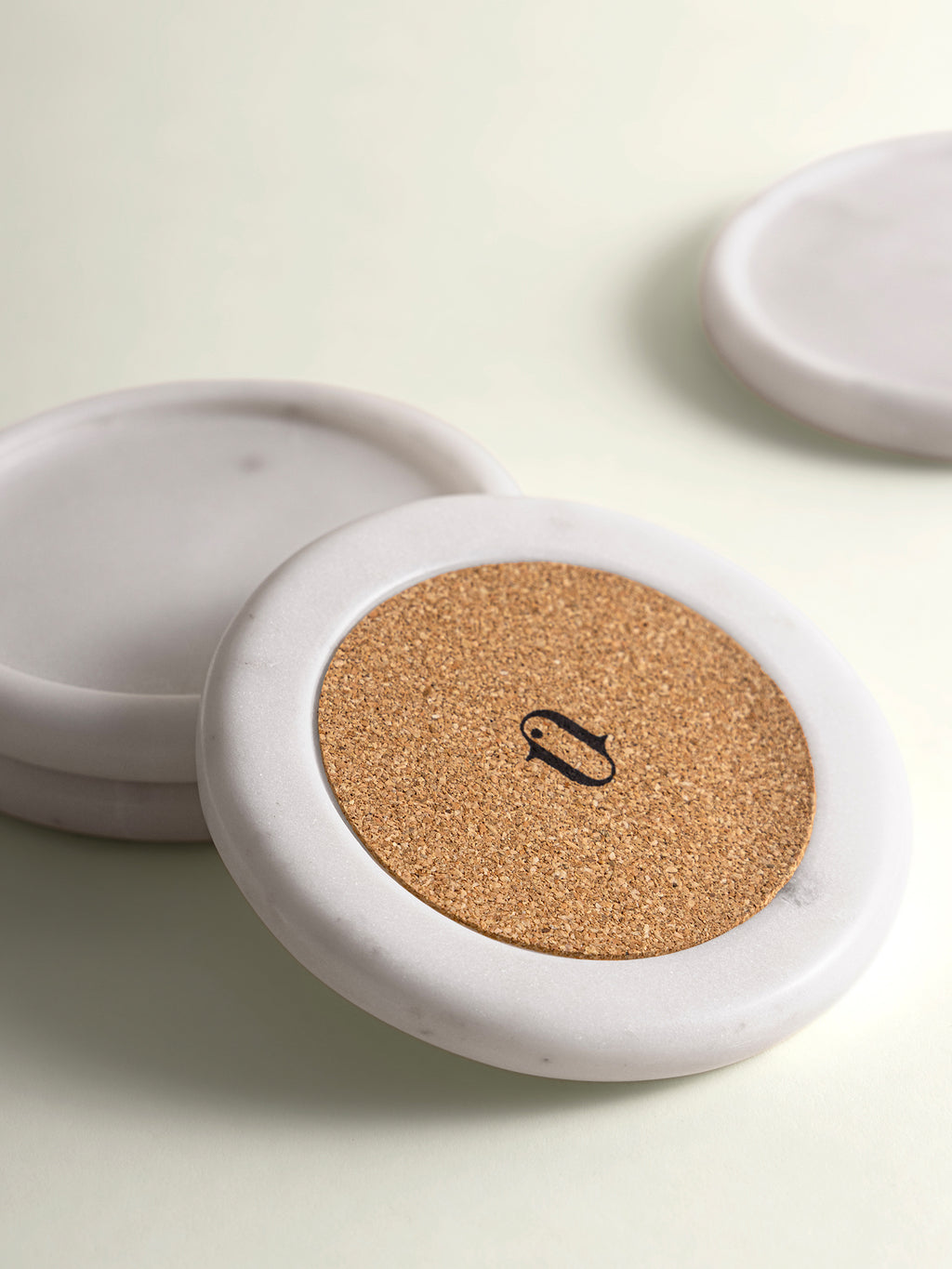 A close-up of a round white marble coaster with a natural cork center with 'anctuar' logo, shown on a light cream background with other coasters blurred in the background.