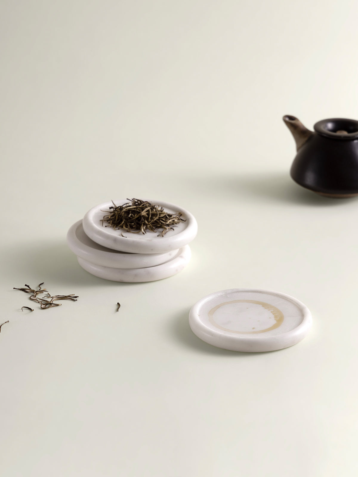 Three small round white marble coasters stacked on a light cream surface, with loose tea leaves on top and one coaster placed in front. A dark teapot is softly blurred in the background.