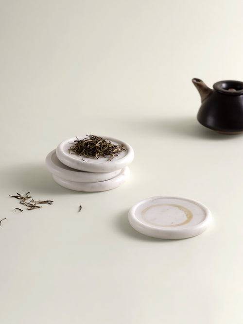 Three small round white marble coasters stacked on a light cream surface, with loose tea leaves on top and one coaster placed in front. A dark teapot is softly blurred in the background.