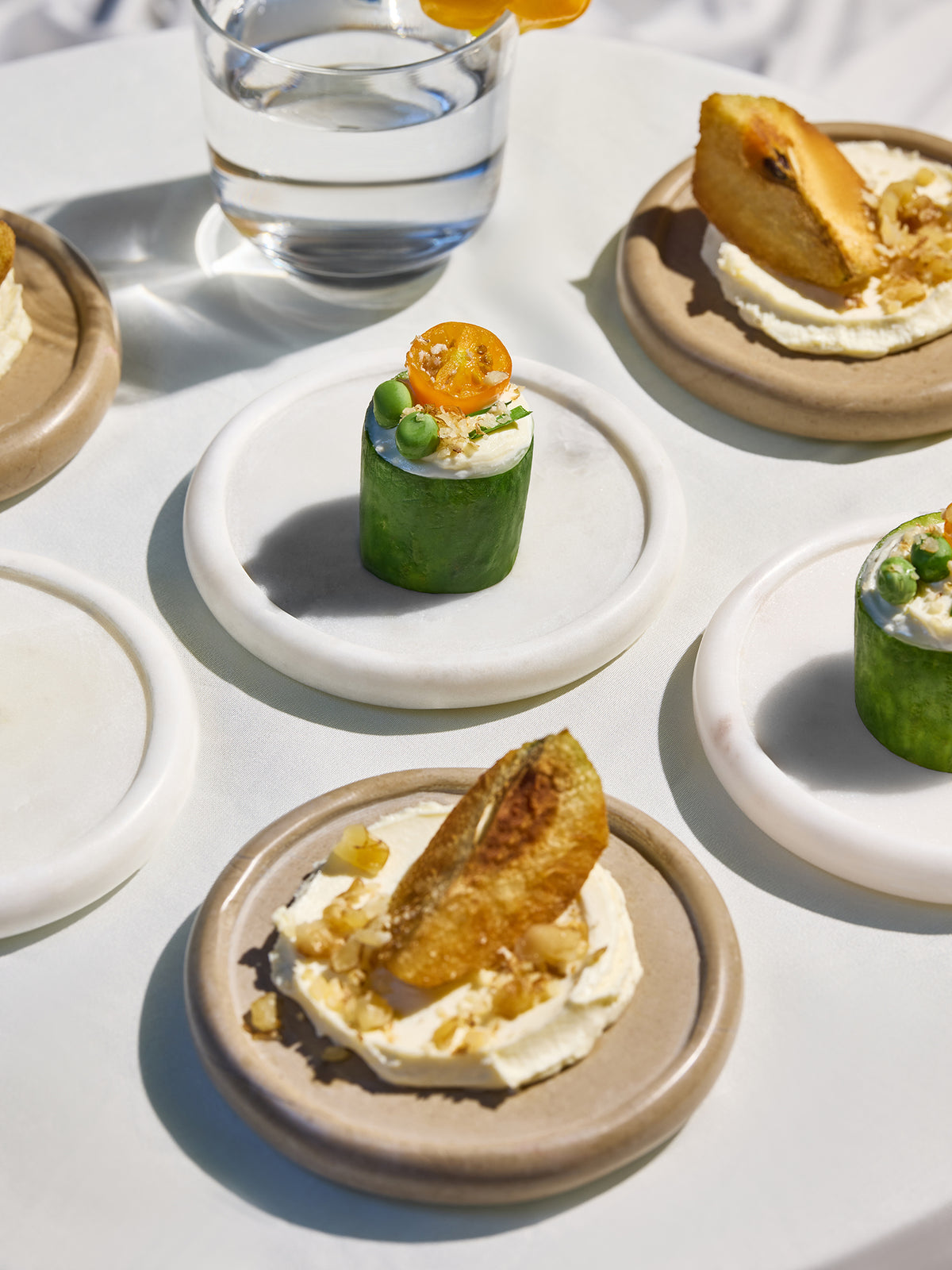 A styled food spread on a white tablecloth with marble coasters, featuring small appetizers including cucumber wraps with cream and orange topping, and topped with crispy fried strips, alongside a glass of water and a yellow chilli garnish.
