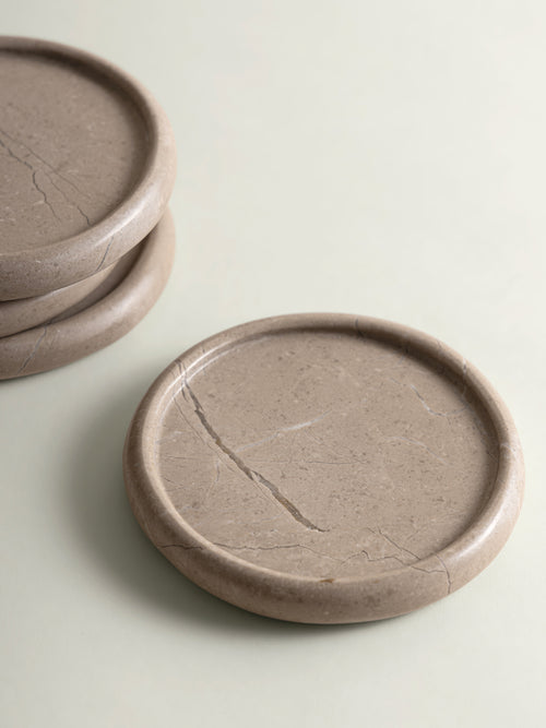 Round marble coasters in a warm taupe-brown color with natural fissures, shown with one stack of two coasters on the left and one single coaster in the foreground on a light cream background.