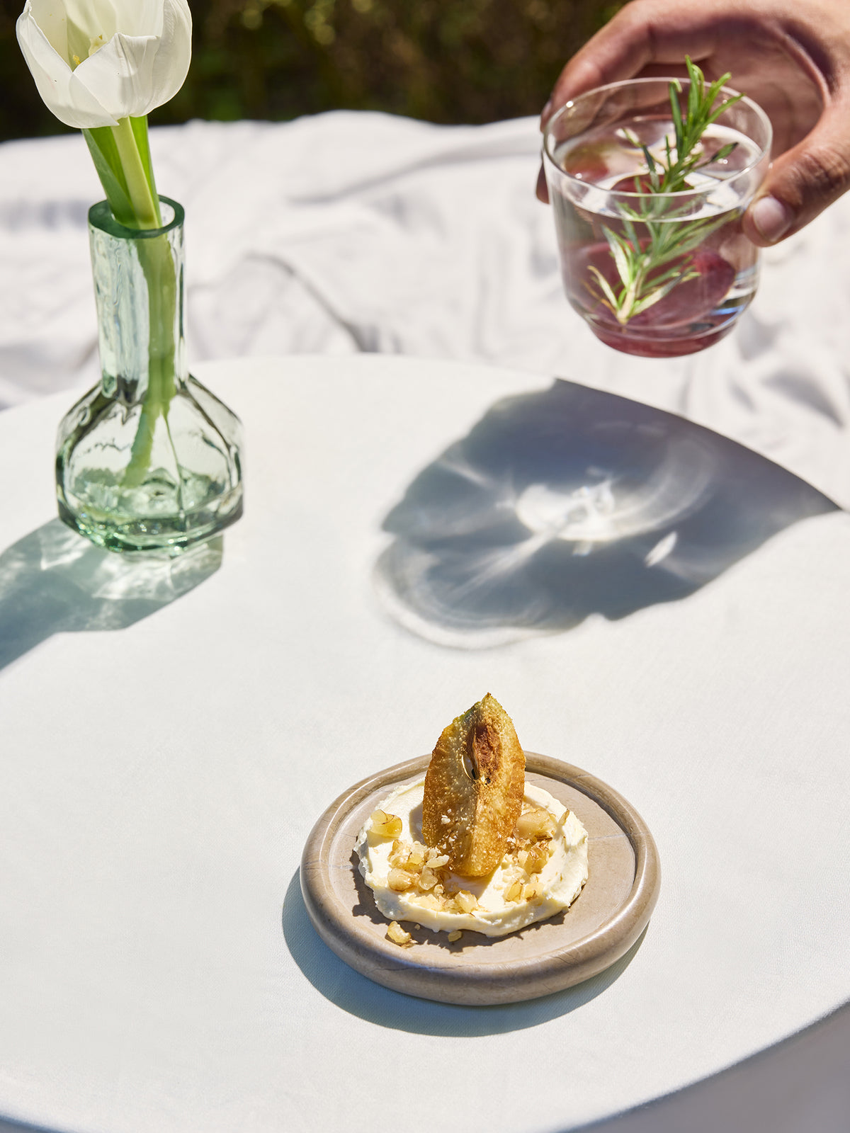 A styled outdoor dining scene with a small plate of dessert on a white tablecloth, a glass vase with a white tulip, and a hand holding a glass of water with a rosemary sprig in the background.