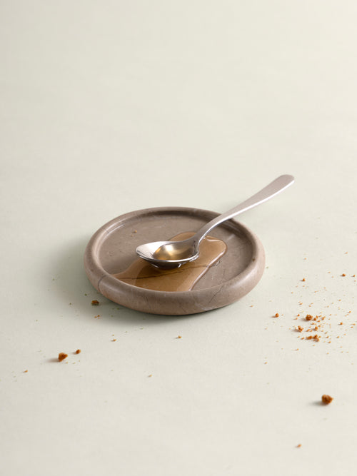 A small round marble coaster in a warm taupe-brown color with a polished silver spoon resting on top, set against a matching light beige background with scattered crumbs.