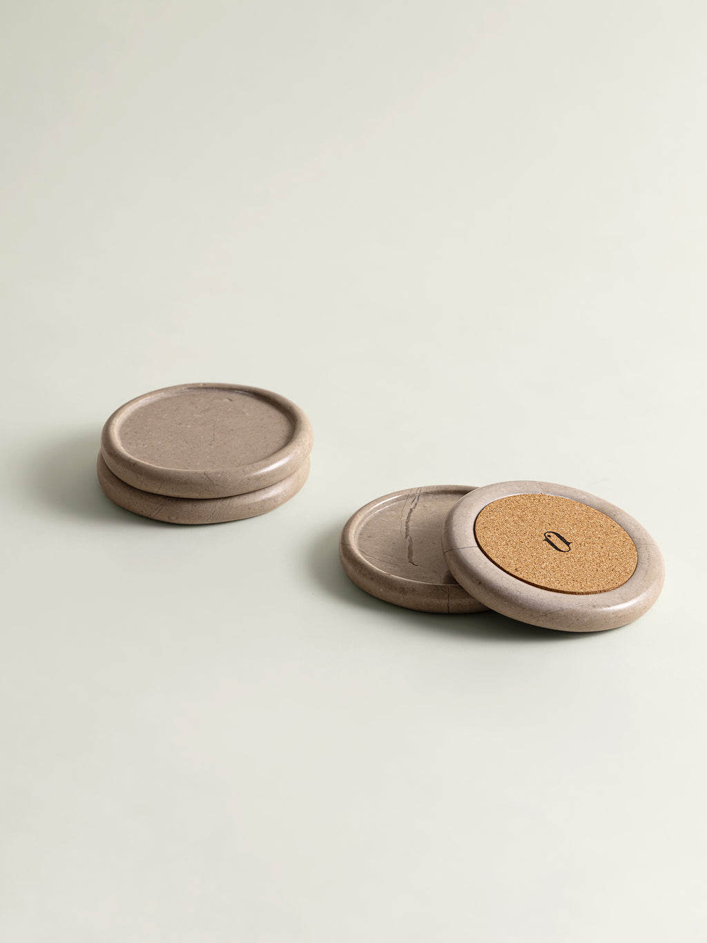 Three round, marble coasters in a warm taupe-beige color, arranged on a light off-white background. One coaster is revealing a textured cork base 