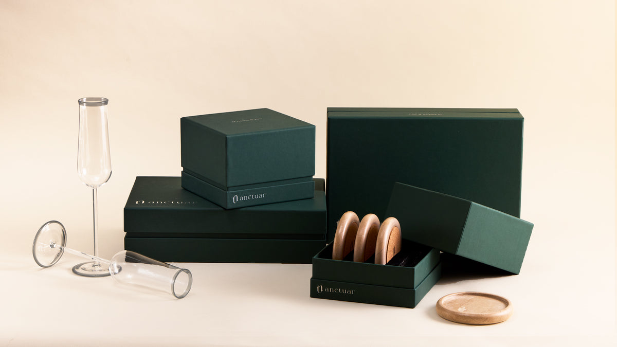 Dark green anctuar gift boxes and packaging displayed on a cream surface, alongside a lipp flute stemmed glass, a small clear tumbler, and a cuar round marble coasters.