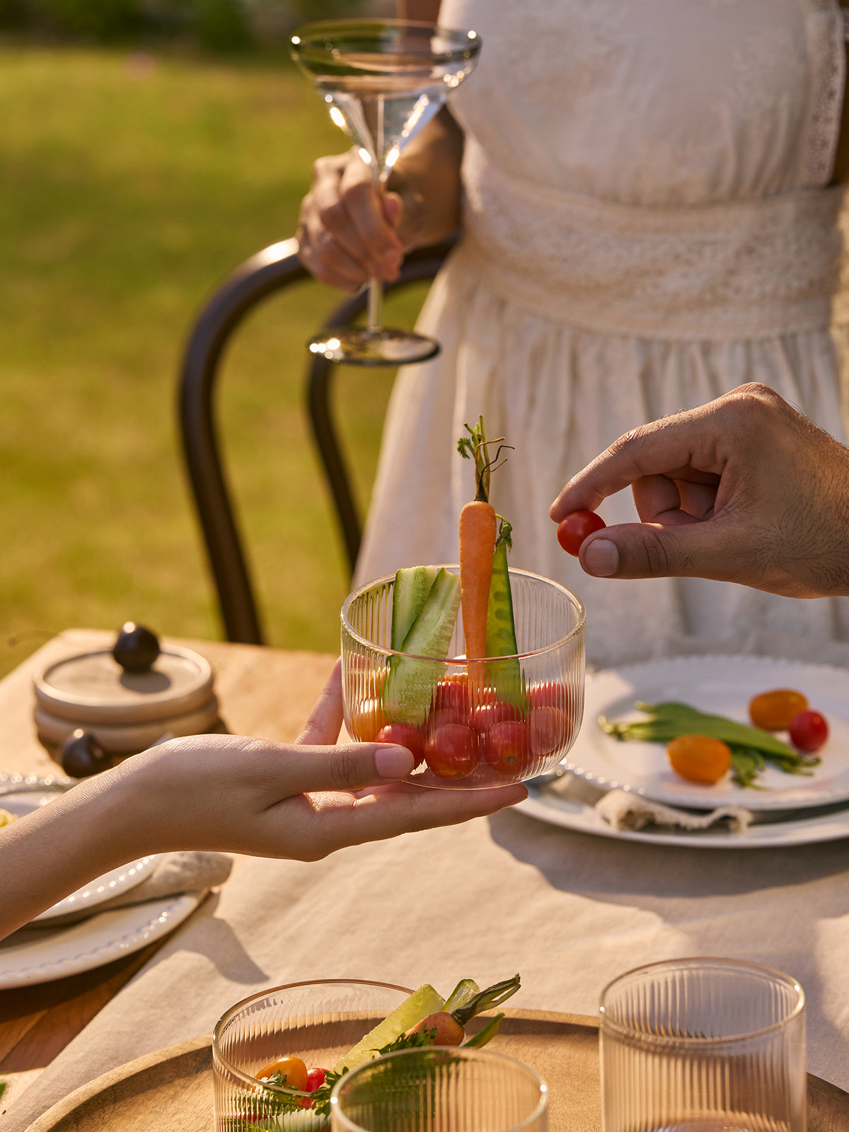 A clear ribbed glass tumbler held by hands at an outdoor dining table, filled with small cherry tomatoes, cucumber sticks, and a carrot garnish. A person in a white dress holds a martini glass in the background, with plates and green grass visible.