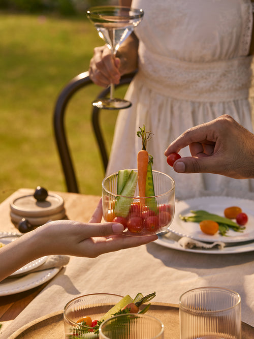 A clear ribbed glass tumbler held by hands at an outdoor dining table, filled with small cherry tomatoes, cucumber sticks, and a carrot garnish. A person in a white dress holds a martini glass in the background, with plates and green grass visible.
