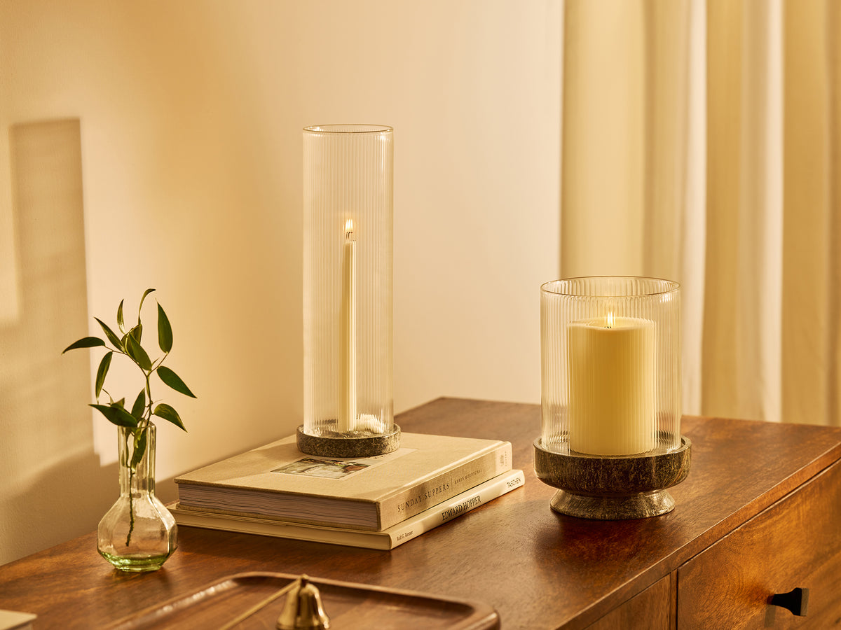 Two tall glass candle holders with white candles displayed on a wooden dresser alongside a small green plant in a glass vase, under warm ambient light.