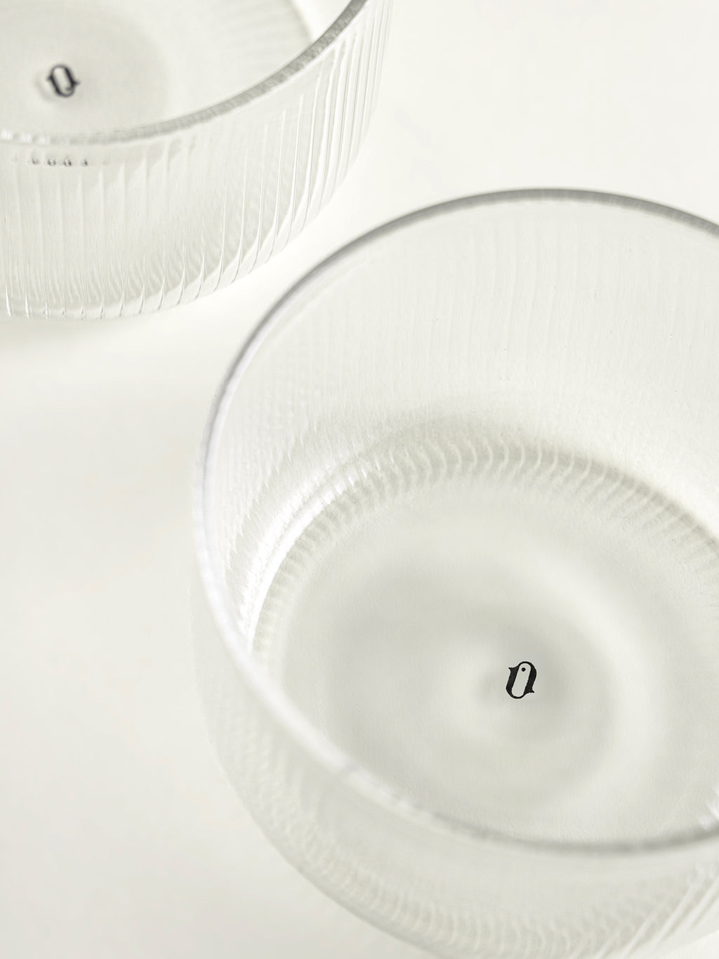Two clear ribbed glass bowls on a white surface, photographed from above with soft lighting and logo printed inside each bowl.