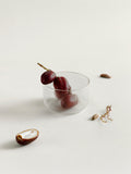 A clear ribbed glass bowl or tumbler with three dark red dates inside, styled on a light beige background with scattered dates and a small dried botanical prop.