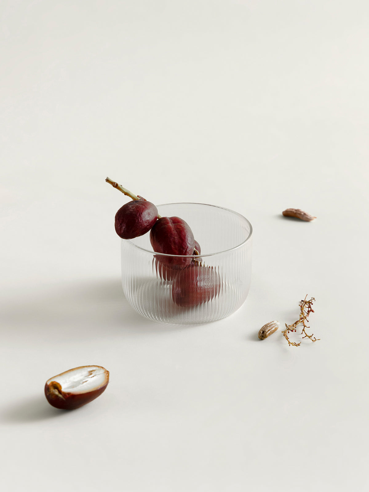 A clear ribbed glass bowl or tumbler with three dark red dates inside, styled on a light beige background with scattered dates and a small dried botanical prop.