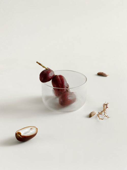 A clear ribbed glass bowl or tumbler with three dark red dates inside, styled on a light beige background with scattered dates and a small dried botanical prop.