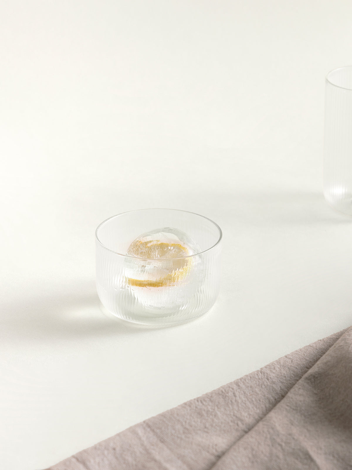 A clear ribbed glass tumbler containing an ice sphere with a slice of orange, placed on a light beige surface with a folded beige cloth in the foreground.