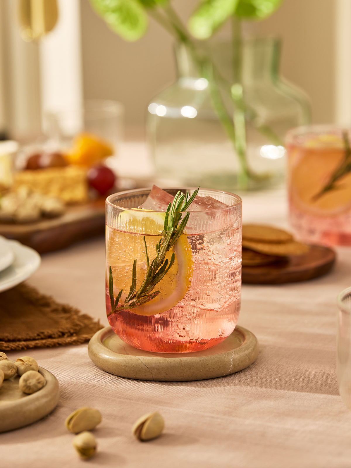 A pink cocktail in a clear ribbed rocks glass with a lemon slice and rosemary sprig, set on a beige coaster on a table with snacks and a blurred background.