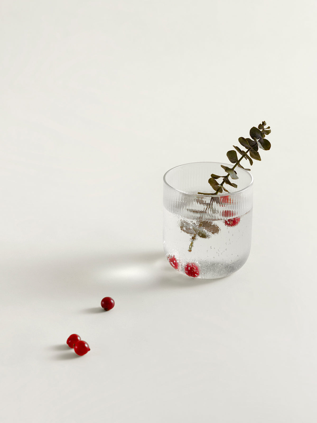 A clear textured glass tumbler containing water, a sprig of green eucalyptus, and small red berries, photographed on a clean off-white background with a few loose red berries nearby.