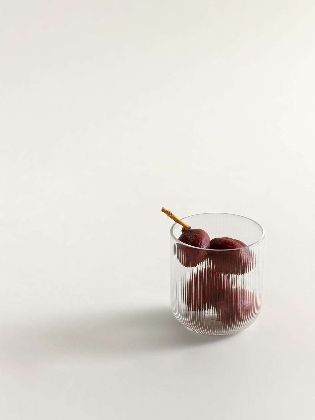A clear ribbed glass tumbler containing dark red dates, photographed on a clean off-white studio background.