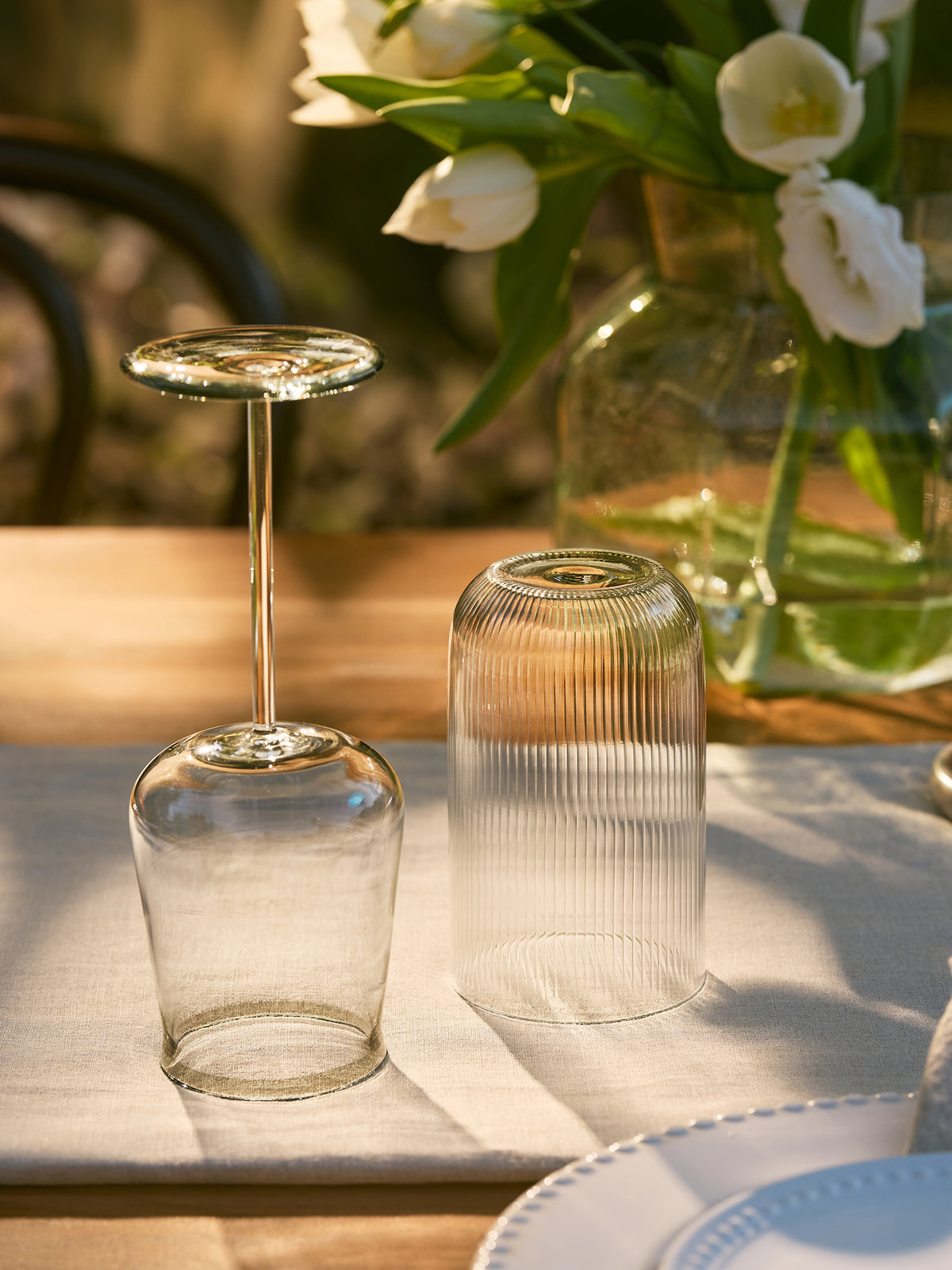 A ribbed glass and a wine glass with rim detail placed on a white tablecloth at a dining table, with a vase of white tulips in the background.