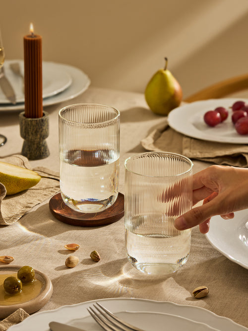 A hand holds a ribbed clear drinking glass on a beige tablecloth in a warm, styled dining setting with plates, a candle, and fruit.