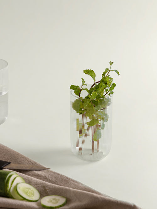 A clear ribbed glass tumbler holding fresh green mint stems, styled on a light neutral surface with cucumber slices and a beige cloth in the foreground.