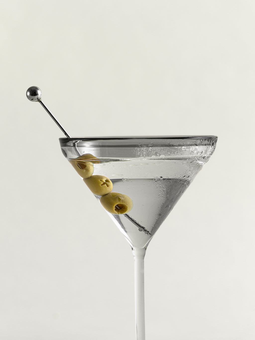 A clear martini glass filled with a transparent cocktail and garnished with three green olives on a metal skewer, photographed against a plain light background.