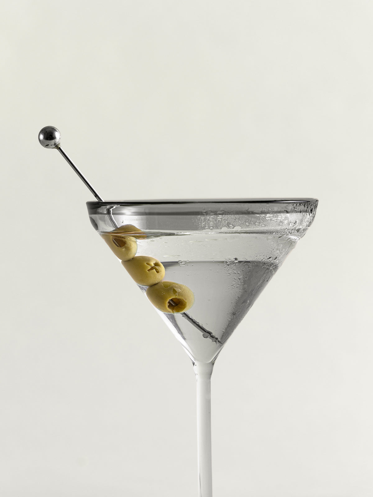A clear martini glass filled with a transparent cocktail and garnished with three green olives on a metal skewer, photographed against a plain light background.