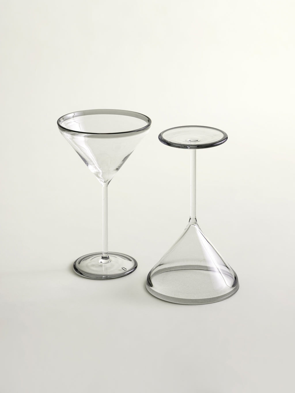 Two clear cocktail glass on a light gray studio background: a martini-style coupe glass and a small conical stemware piece with a grey circular rim.