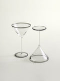 Two clear cocktail glass on a light gray studio background: a martini-style coupe glass and a small conical stemware piece with a grey circular rim.