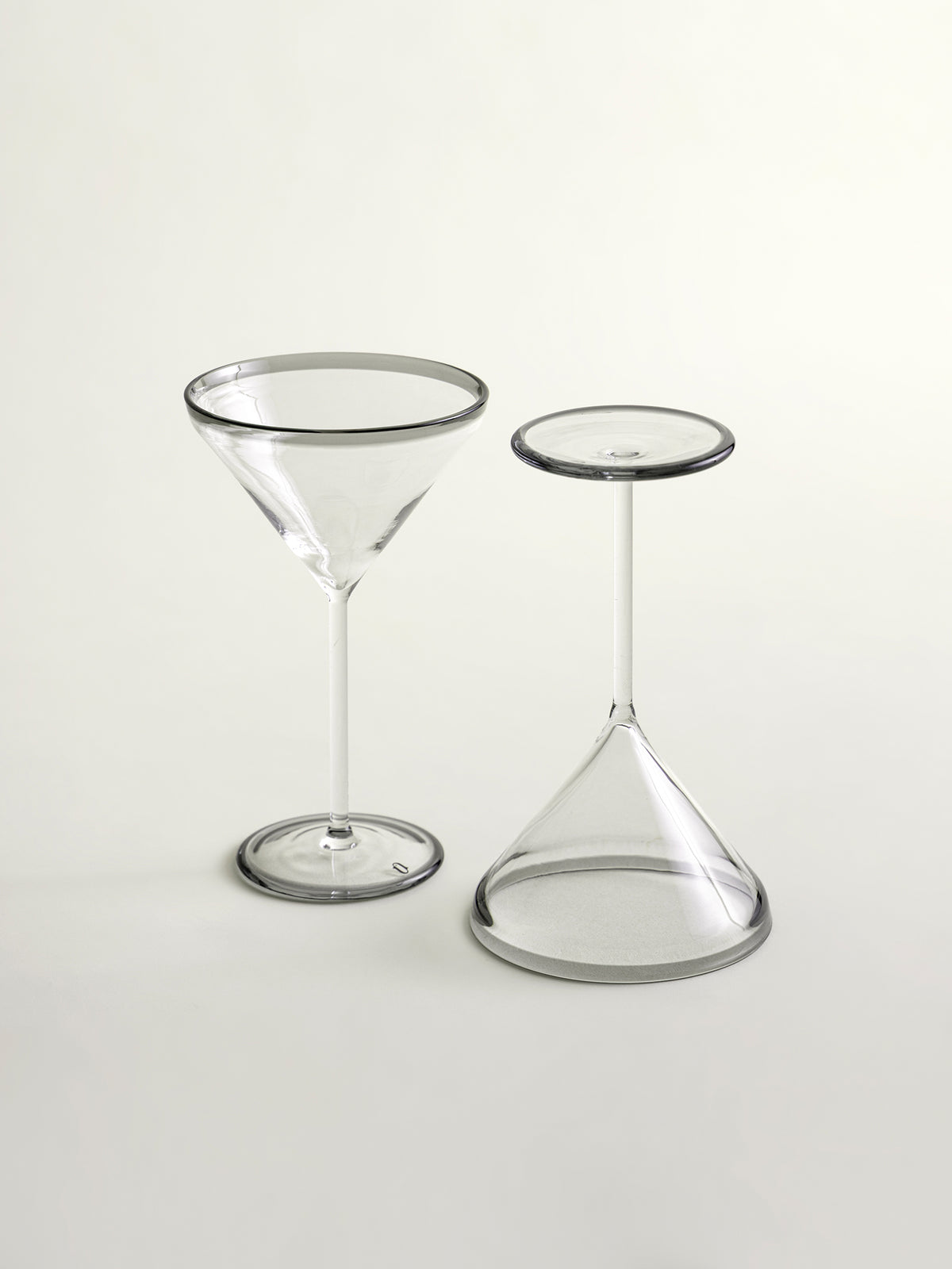 Two clear cocktail glass on a light gray studio background: a martini-style coupe glass and a small conical stemware piece with a grey circular rim.