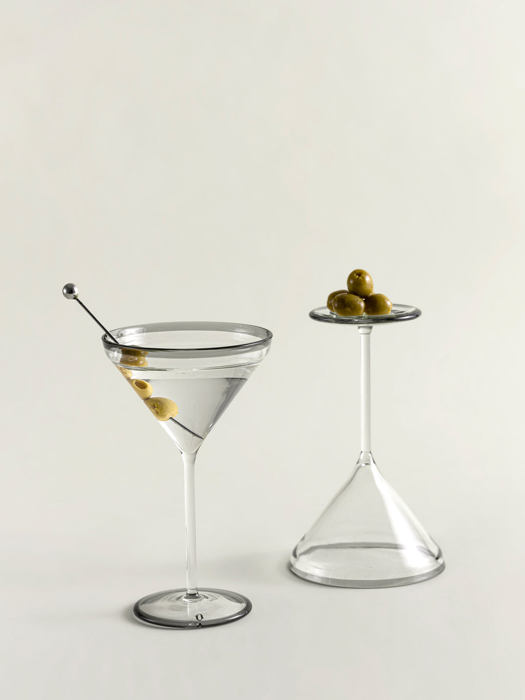 Two clear glass cocktail-style glasses on a light neutral background, one filled with a clear drink and one empty, each with a small cluster of green olives on a glass garnish.