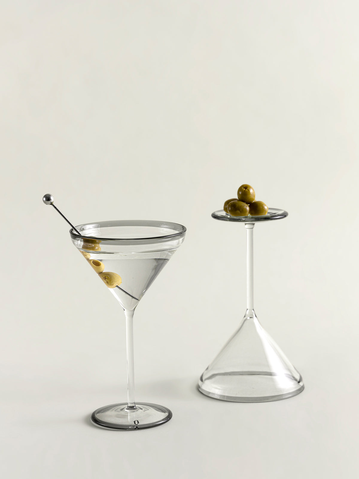 Two clear glass cocktail-style glasses on a light neutral background, one filled with a clear drink and one empty, each with a small cluster of green olives on a glass garnish.