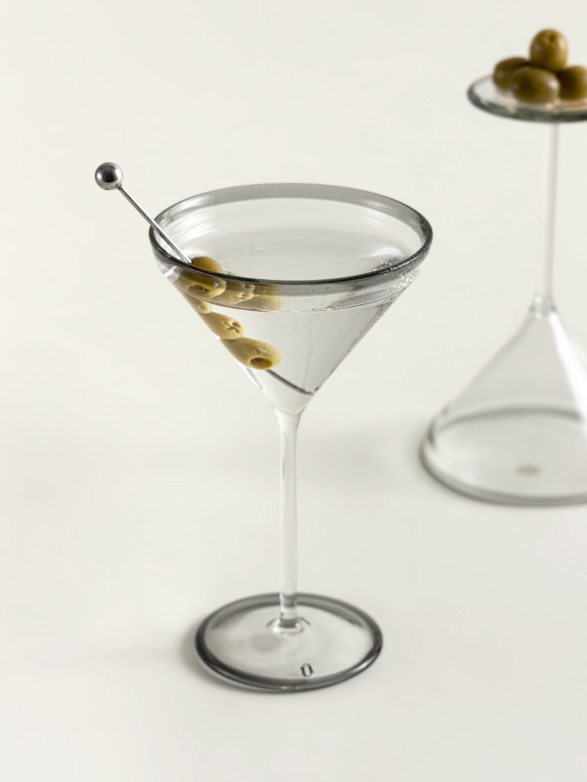 A clear martini glass filled with a transparent liquid and garnished with green olives on a metal skewer, photographed on a clean white background with a softly blurred second glass in the background.