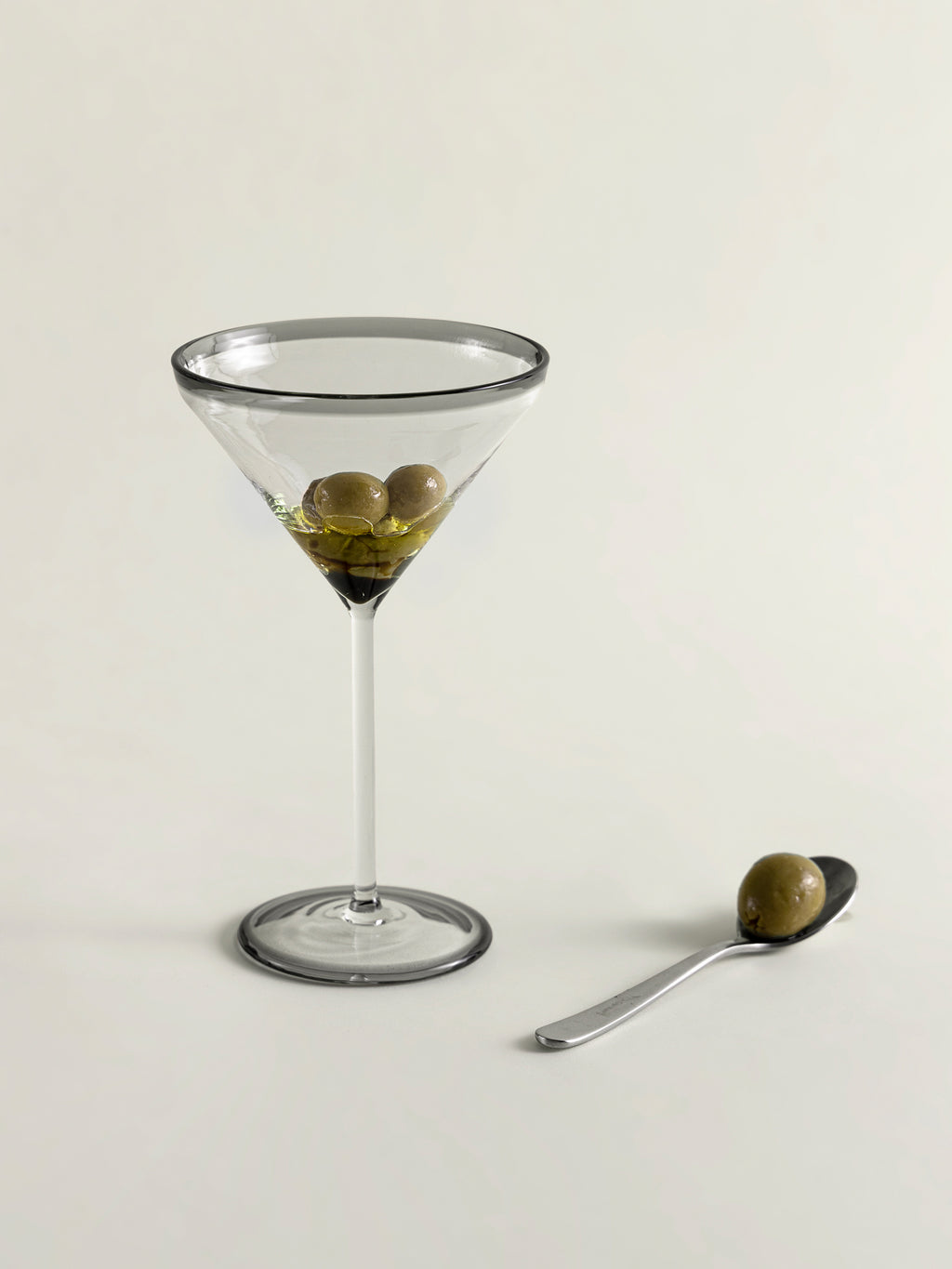 A clear glass martini glass with smoke grey rim and three green olives in olive oil, shown on a light beige background next to a small spoon holding one olive.