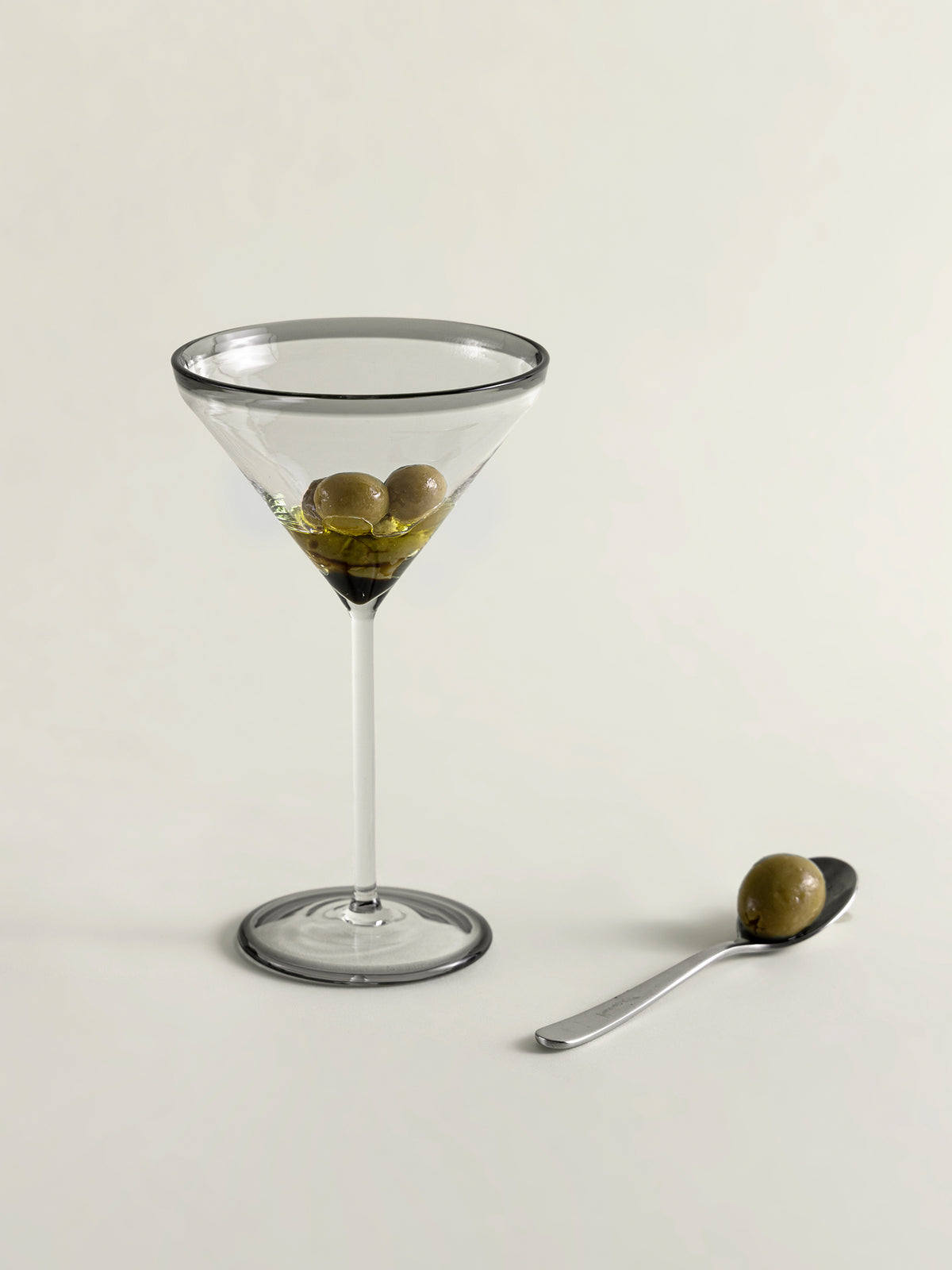 A clear glass martini glass with smoke grey rim and three green olives in olive oil, shown on a light beige background next to a small spoon holding one olive.