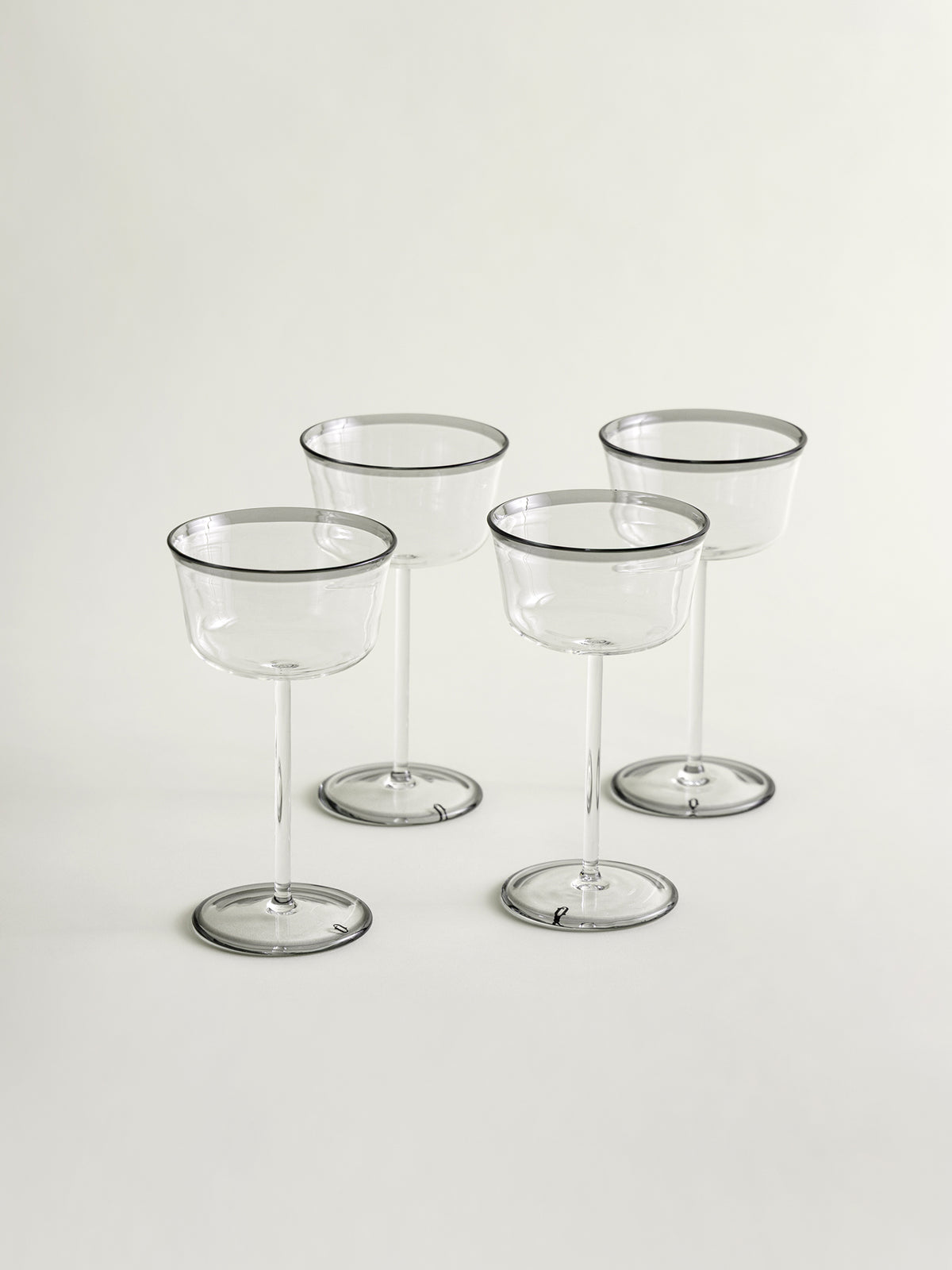 Four clear stemmed coupes with thin smoke grey rims, arranged on a light neutral background.