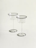 Two clear stemmed coupes with thin smoke grey rims, standing on a light neutral background.