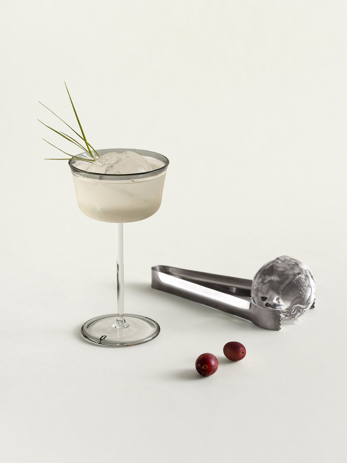 A coupe glass with a green garnish, shown beside a metal ice ball maker and two cranberries on a clean off-white background.