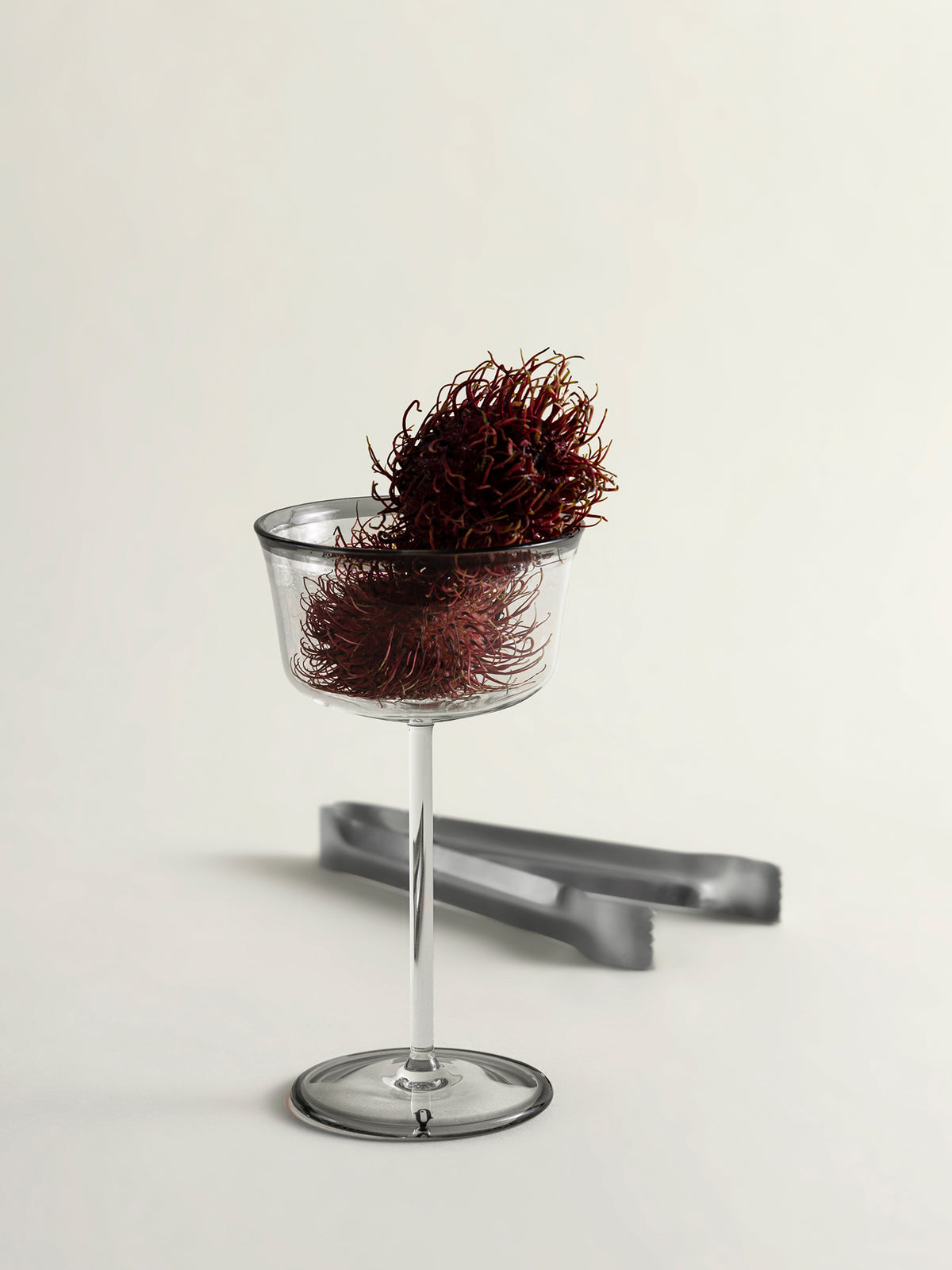A clear stemmed coupe glass with a dark red rambutan inside and resting on top, shown on a clean off-white studio background with a pair of gray tongs behind it.