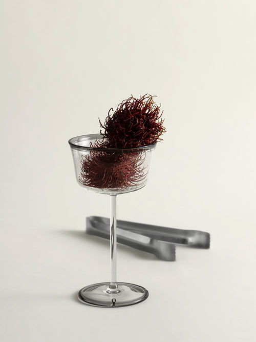 A clear stemmed coupe glass with a dark red rambutan inside and resting on top, shown on a clean off-white studio background with a pair of gray tongs behind it.