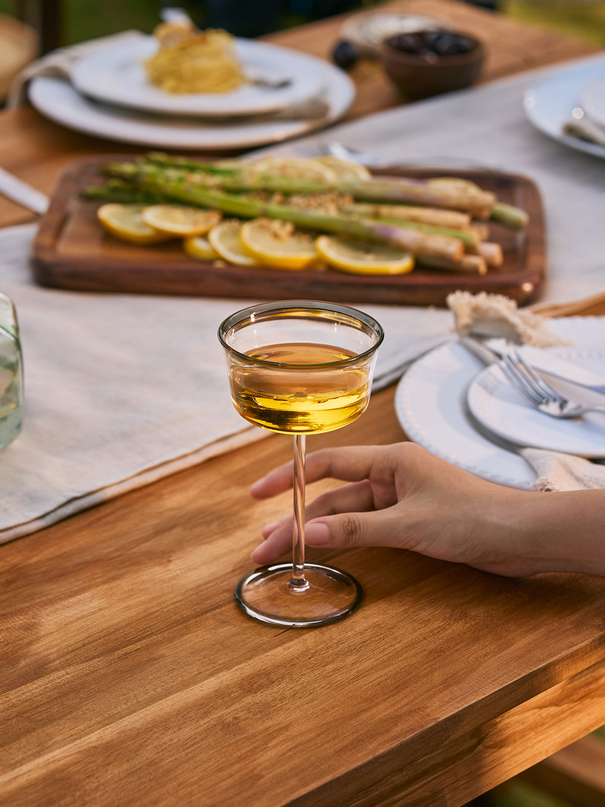 A clear coupe glass with smoke rim detail with a golden yellow drink, held by a person's hand on a wooden dining table with plates, asparagus, and lemon slices in the background.