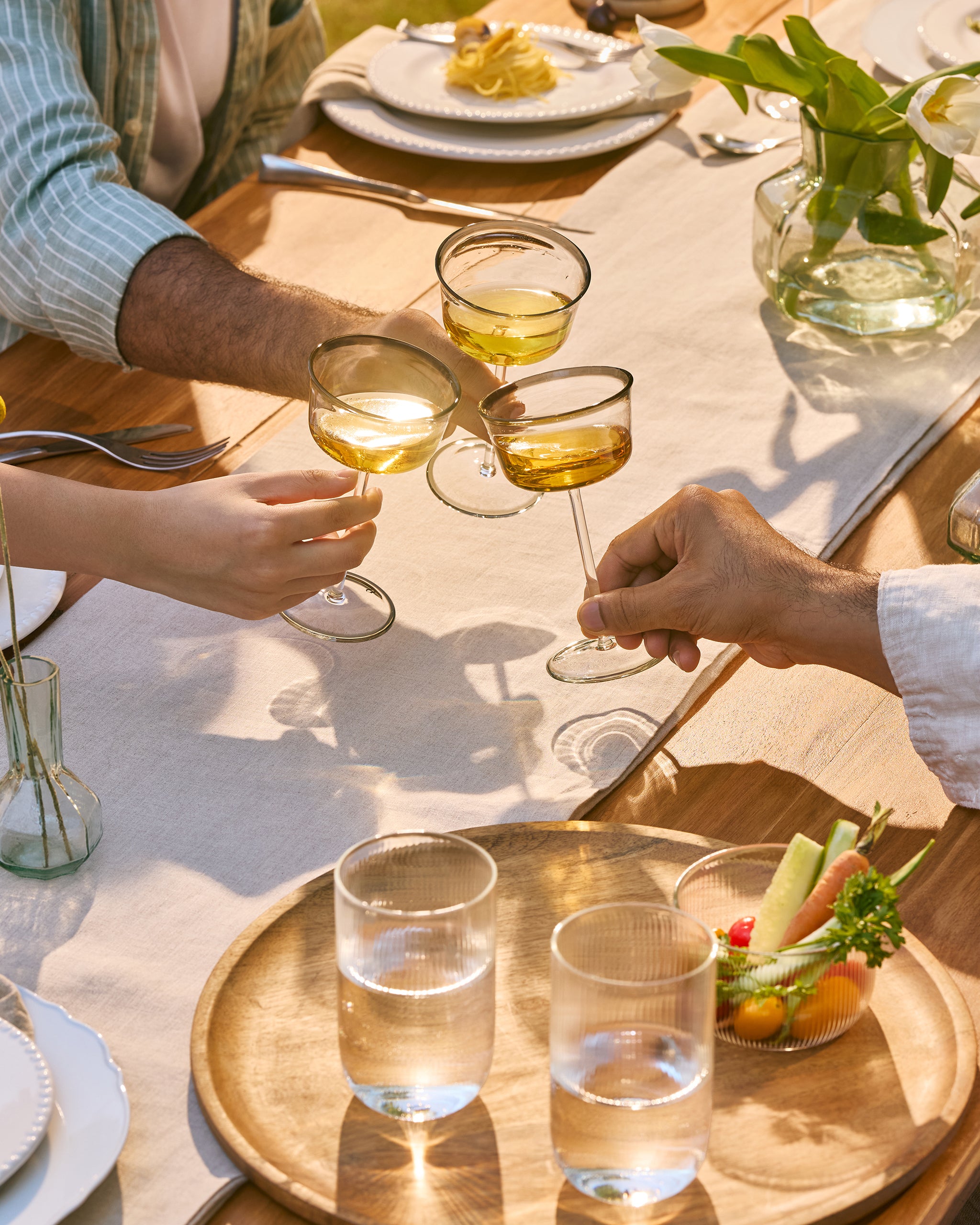 A lifestyle dining scene featuring three people sitting around a wooden table outdoors, clinking coupe glasses filled with white wine. The table is decorated with a white runner, plates, a glass vase with white flowers, and a wooden serving tray holding two glasses of water and a small fruit bowl.